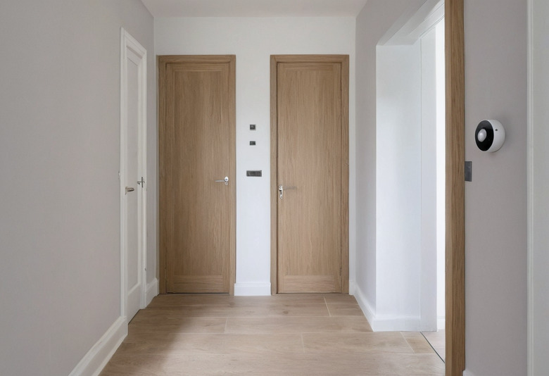 A hallway in an English house with two oak veneer doors side by side, a third oak-lined opening to the right, a pale stone floor and a round wall control beside the opening.