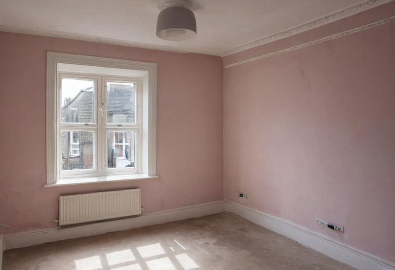 An empty room in an English house with dusky pink walls, a white-framed window, a panel radiator beneath it, a ceiling pendant and a bare subfloor awaiting its finish.