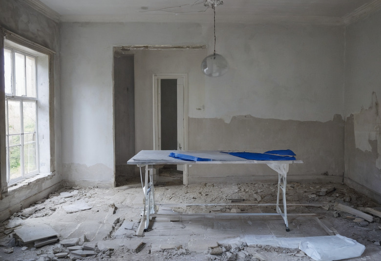 A large room under renovation with plaster removed to waist height, a paste table carrying blue sheeting in the middle of the floor, and broken plaster and dust sheets around it.
