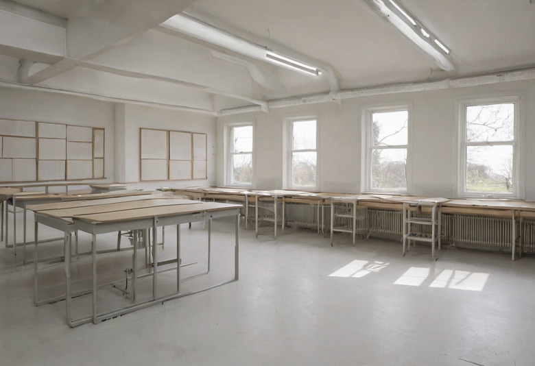 An empty teaching studio with rows of trestle tables along a window wall, cast iron radiators beneath the windows, strip lights on a white vaulted ceiling and blank boards on the end wall.
