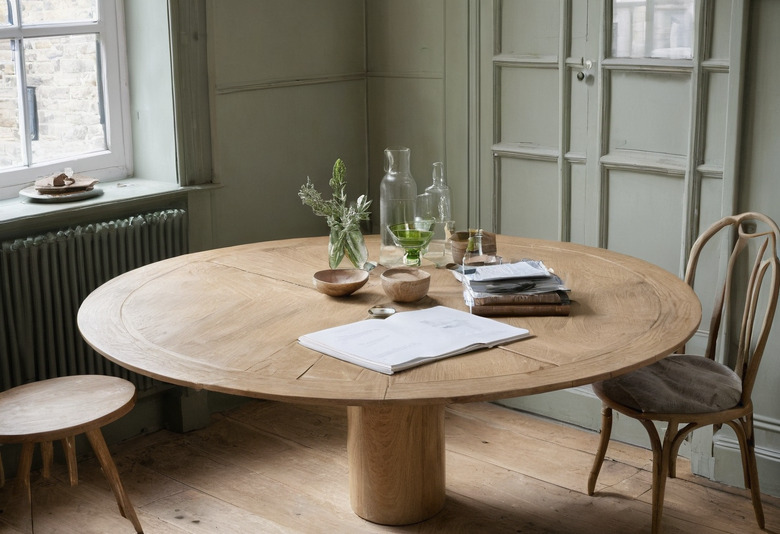 A round oak table in a sage-green panelled room with a column radiator under the window, set with an open document, a stack of books, glass carafes and small wooden bowls.