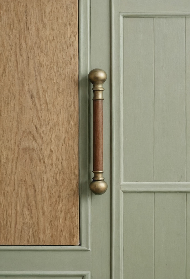 A close view of a cabinet front in plain oak beside a sage-green painted panel, with a long antique brass bar handle mounted between them.