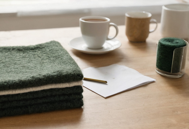 A pale wood table by a window with a folded stack of green and cream wool throws, a cup of tea, a stoneware mug, a rolled wool sample in a glass and a blank sheet with a brass pencil.