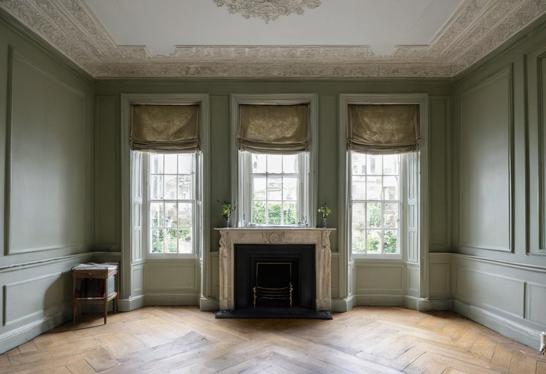 A panelled drawing room painted sage green with an enriched plaster cornice, three sash windows in a canted bay, a stone chimneypiece with a cast iron grate and a herringbone parquet floor.