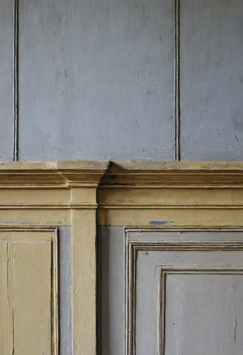 A close view of old painted panelling where a moulded rail in ochre meets grey-blue panels above and below, the paint layers thick and cracked.