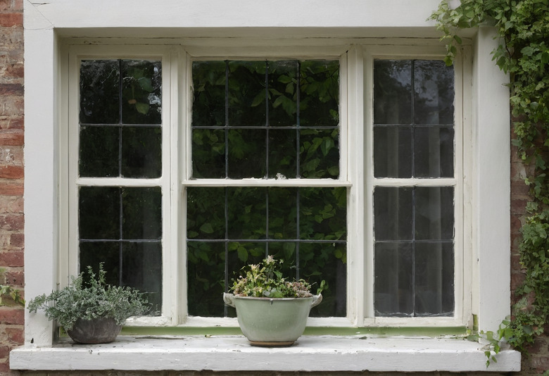 A white-painted timber window with small leaded panes set in a red brick wall, seen from outside, with ivy at one side and two planted pots on the sill.